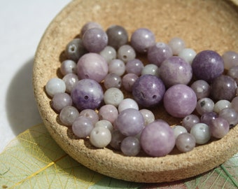 May include: A bowl of purple and white gemstone beads. The beads are round and smooth, and they are arranged in a random pattern. The bowl is made of cork and has a natural finish.
