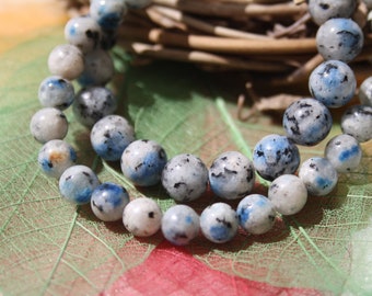 May include: A strand of blue and white gemstone beads. The beads are round and have a speckled pattern. The beads are strung on a clear string and are lying on a green leaf.