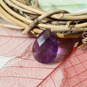May include: A purple glass teardrop-shaped crystal pendant with a faceted design. The pendant is resting on a delicate, red, leaf-like surface.