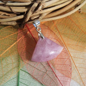 Peut inclure: Un pendentif en pierre précieuse triangulaire, de couleur violet clair, avec une bélière en argent. Le pendentif est posé sur un fond de feuilles colorées et d'un élément de brindilles tressées.