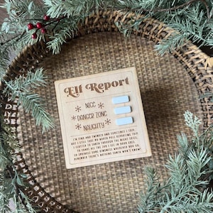 Op de afbeelding: Een houten "Elf Report" bord met de woorden "Nice", "Danger Zone" en "Naughty" en een gedicht. Het bord is omgeven door een gevlochten mand en groenblijvende takken met rode bessen, wat een feestelijk vakantiethema creëert.