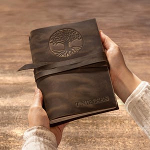 Peut inclure: Un carnet en cuir marron foncé avec un emblème d'arbre de vie en relief sur la couverture avant et une fermeture à lanière en cuir. Le nom T. NIELSEN est gravé dans le coin inférieur droit de la couverture, qui présente une finition mate lisse.