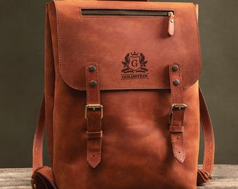 Könnte beinhalten: Ein brauner Lederrucksack mit Klappenverschluss, einem oberen Griff und verstellbaren Schultergurten. Der Rucksack hat eine Reißverschlusstasche vorne und zwei Schnallenverschlüsse. Das Logo "GOLDSTEIN" ist auf der Vorderseite geprägt.