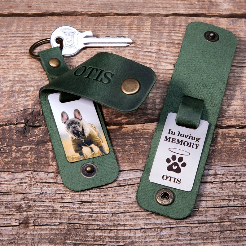 May include: Two dark green leather keychains with brass snap buttons. One keychain holds a metal tag with a photo of a French Bulldog and the name OTIS embossed on the leather. The other tag features the text In loving MEMORY with a paw print.