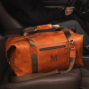 May include: A tan leather duffel bag featuring a brown nylon strap, a zippered front pocket, and a personalised leather handle wrap embossed with the name MANDY. The bag displays a large letter M with a floral design on the front panel.