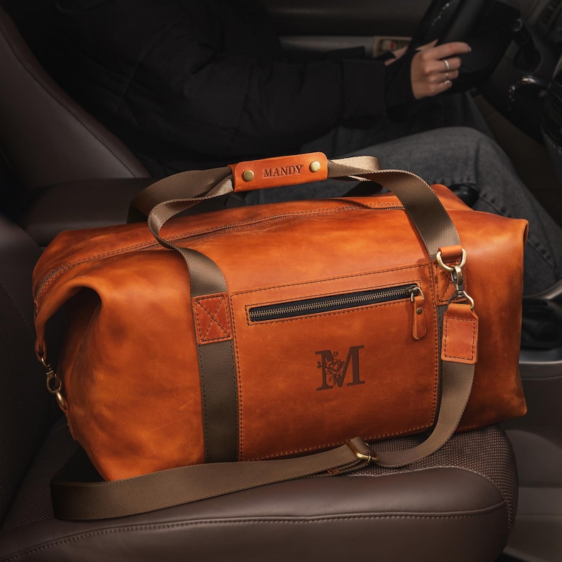 May include: A tan leather duffel bag featuring a brown nylon strap, a zippered front pocket, and a personalised leather handle wrap embossed with the name MANDY. The bag displays a large letter M with a floral design on the front panel.