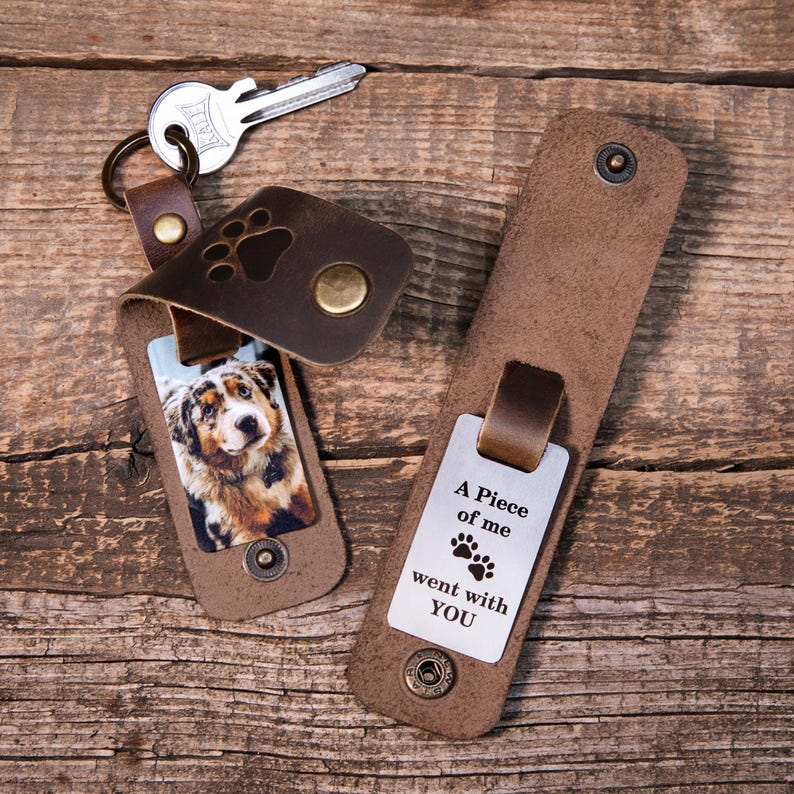 May include: Two brown leather keychains with brass snaps. One keychain features a photo of a dog and a paw print cutout. The other keychain displays a metal plate engraved with the text A Piece of me went with YOU and two paw prints.