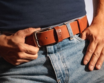 May include: A tan leather belt with a matte silver-toned metal buckle and a leather keeper loop embossed with the text AMIK. The belt is worn through the belt loops of light blue denim jeans, secured over a dark navy blue t-shirt.