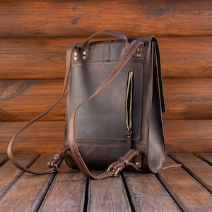 May include: A dark brown leather backpack featuring a vertical zippered pocket on the back panel, adjustable shoulder straps with brass-toned metal buckles, a top carry handle, and reinforced stitching with visible metal rivets at the strap attachments.