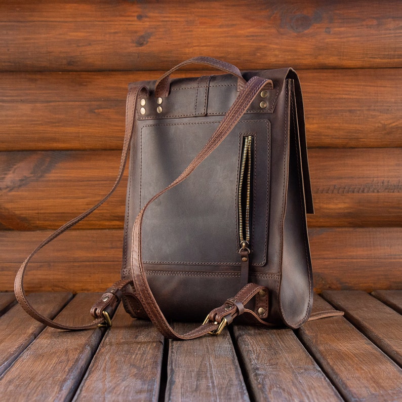 May include: A dark brown leather backpack featuring a vertical zippered pocket on the back panel, adjustable shoulder straps with brass-toned metal buckles, a top carry handle, and reinforced stitching with visible metal rivets at the strap attachments.