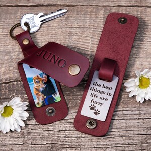 May include: Two burgundy leather keychains with brass snap closures. One keychain features the name JUNO embossed on the leather and a photo of a person with a dog. The other keychain displays the text the best things in life are furry.