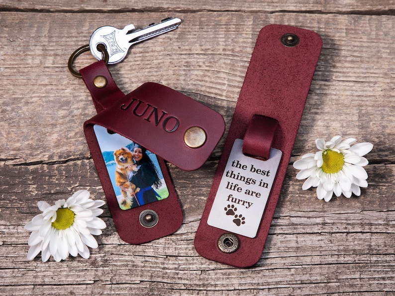 May include: Two burgundy leather keychains with brass snap closures. One keychain features the name JUNO embossed on the leather and a photo of a person with a dog. The other keychain displays the text the best things in life are furry.