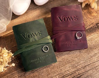 May include: Two leather vow books, one dark green and one deep burgundy, each featuring a wrap-around leather tie, the embossed word VOWS, and the names LUCAS & AMELIA with dates 6.22.2024 and 6.23.2024. A silver band rests on each book cover.