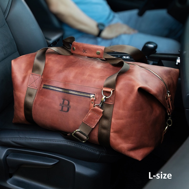 May include: A tan leather holdall with brown nylon straps, a top handle featuring the initials BC, and a front pocket displaying the letter B and the name Brandon. The bag includes a detachable shoulder strap and is labelled as L-size in the corner.