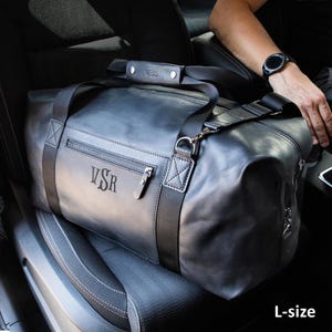 May include: A large charcoal grey leather duffel bag featuring black nylon webbing straps, a zippered front pocket, and a leather handle wrap. The bag is embossed with the letters VSR on the front pocket and the handle wrap. The text L-size is visible.