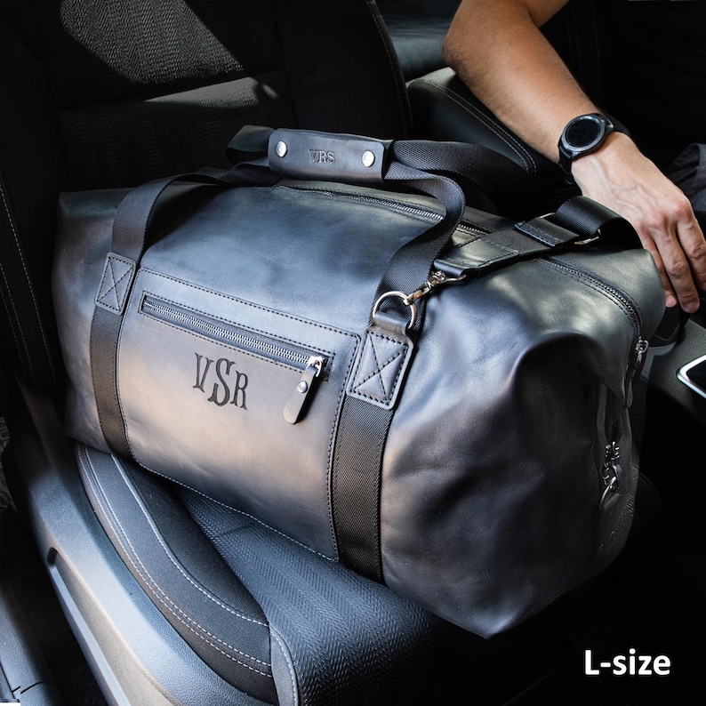 May include: A large charcoal grey leather duffel bag featuring black nylon webbing straps, a zippered front pocket, and a leather handle wrap. The bag is embossed with the letters VSR on the front pocket and the handle wrap. The text L-size is visible.