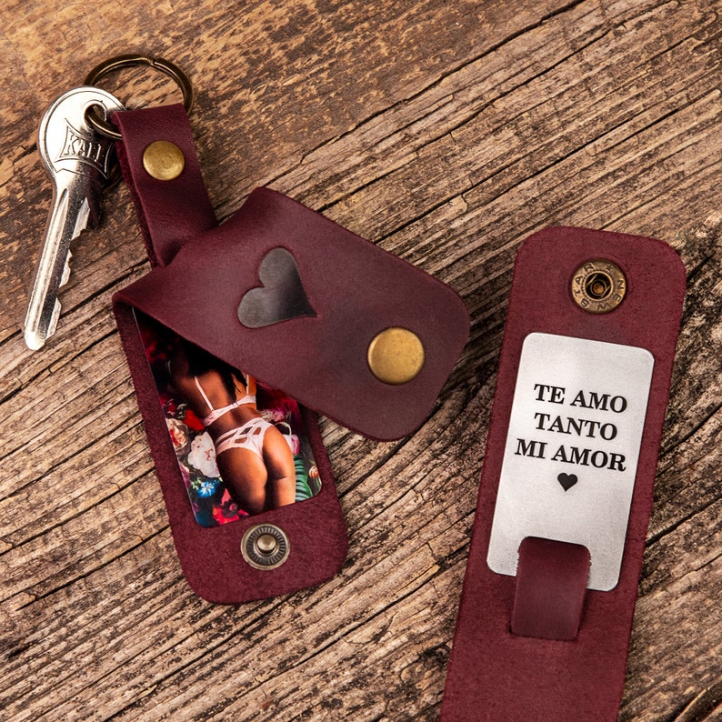 May include: A burgundy leather keychain with a heart-shaped cutout, a metal snap closure, and a metal key ring. The interior displays a photograph of a person in white lingerie. A second leather piece features a metal plate engraved with TE AMO TANTO MI AMOR.