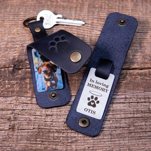 May include: A dark blue leather keychain with a metal snap closure and a debossed paw print. One side features a photo of a brown dachshund, while the other displays a metal tag engraved with "In loving MEMORY" above a paw print and "OTIS".