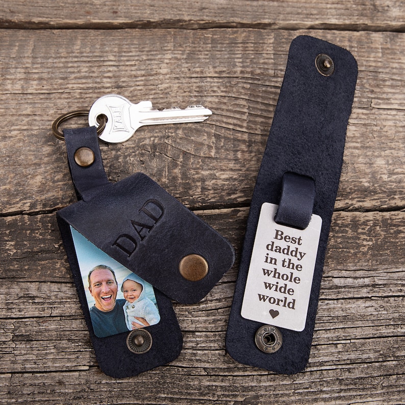 May include: Two dark blue leather key holders with brass snaps. One holder is open, revealing a photo of a man and a child, with the word DAD embossed on the flap. The other holder is closed, displaying a metal tag engraved with Best daddy in the whole wide world and a heart symbol.