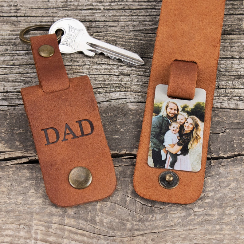May include: Two brown leather keychains with antique brass hardware. One keychain features the word DAD embossed in black, while the other holds a rectangular metal insert displaying a color photograph of a family of four. A silver key is attached.