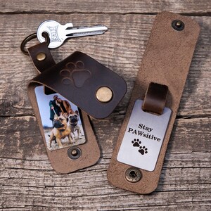 May include: Two dark brown leather keychains with brass snap closures. One keychain holds a metal key and features a photo of two dogs and a person. The other keychain is open, revealing a silver metal tag engraved with the text Stay PAWsitive.
