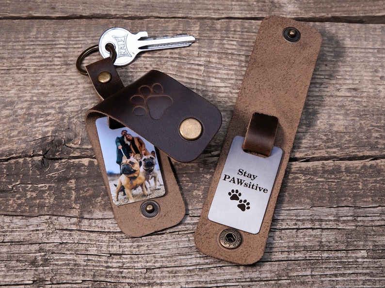 May include: Two dark brown leather keychains with brass snap closures. One keychain holds a metal key and features a photo of two dogs and a person. The other keychain is open, revealing a silver metal tag engraved with the text Stay PAWsitive.