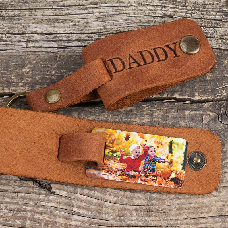 May include: A brown leather keychain featuring the word DADDY embossed on the top flap. The lower section includes a rectangular metal insert displaying a color photograph of two children sitting in a pile of autumn leaves, secured by a metal snap.