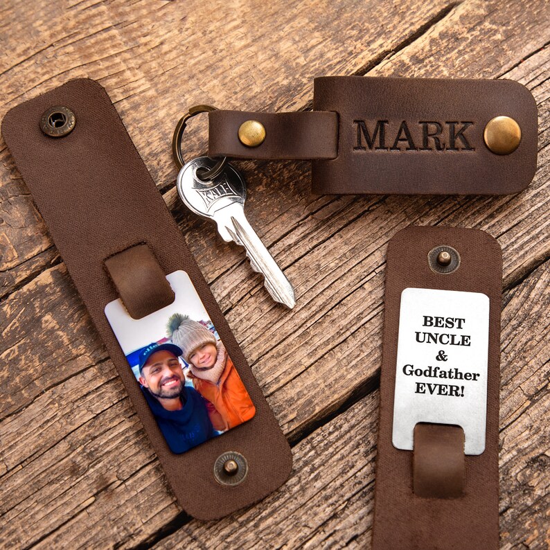 May include: Dark brown leather keychains with brass snaps. One features the name MARK embossed, another holds a metal plate with a photo of two people, and a third displays the engraved text BEST UNCLE & GODFATHER EVER! with a metal key.