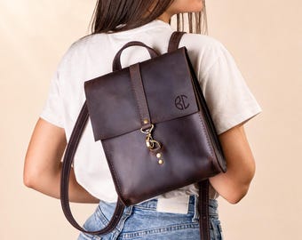 May include: A dark brown leather backpack featuring a fold-over flap with a brass-toned metal clasp closure. The front of the flap displays a circular monogram with the letters BC. The bag includes thin leather shoulder straps and a top handle.