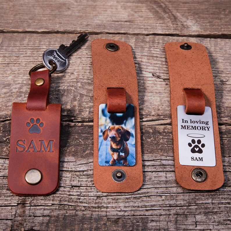 May include: Three brown leather keychains with metal snaps. One features a stamped paw print and the name SAM. Another displays a photo of a brown dachshund. The third shows a metal tag with the text In loving MEMORY, a halo, and a paw print.