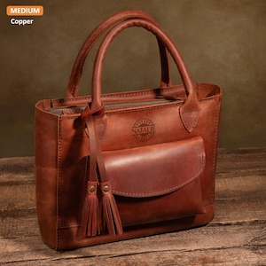 May include: A brown leather tote bag with a zipper closure and a front pocket. The bag has a tassel detail and the name "Natalie" is embossed on the front.