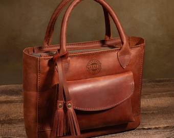 May include: A brown leather tote bag with a zip closure and a front pocket. The bag has a tassel detail and the name "Natalie" is embossed on the front.