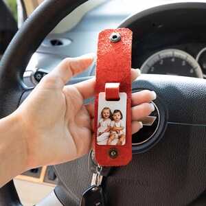 May include: A red textured leather key fob strap with a metal snap button closure. A rectangular white tag featuring a printed photograph of two young children is attached to the strap. A metal key ring is connected to the bottom of the leather strap.