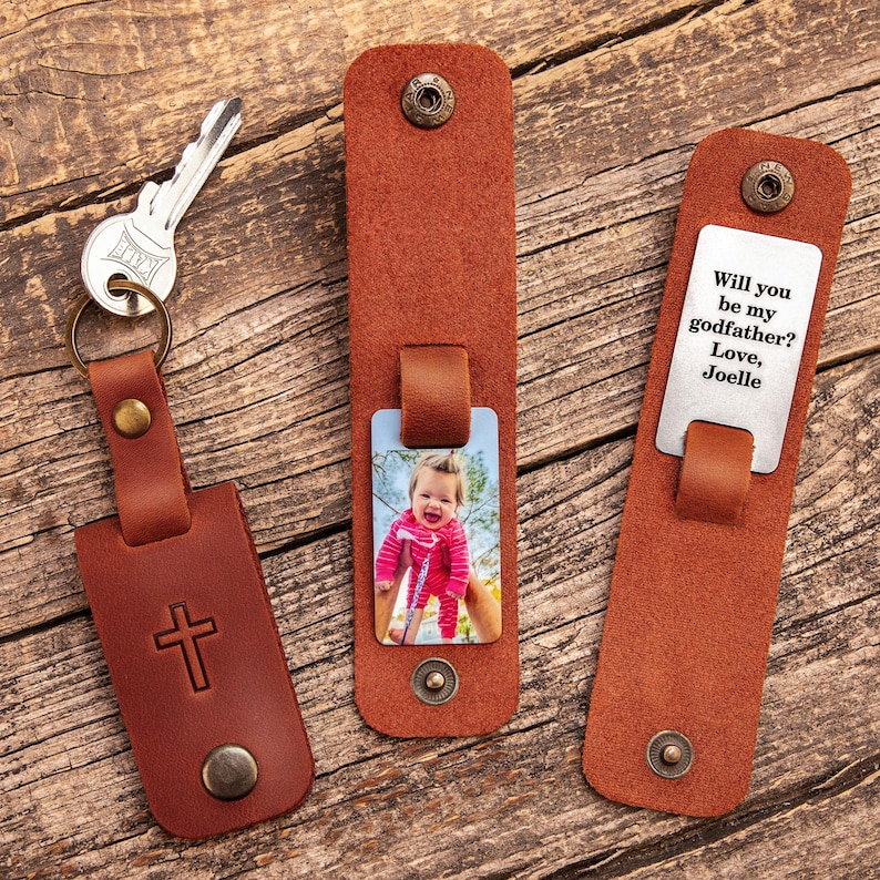 May include: Three brown leather keychains with metal snaps. One features a cross emblem, one displays a photo of a child in a pink outfit, and one shows the text: Will you be my godfather? Love, Joelle. A silver key is attached to the cross keychain.