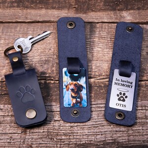 May include: Three dark blue leather key organizers with metal snaps. One features a paw print embossment and a silver key. Another displays a photo of a brown dachshund. The third shows a metal tag with the text In loving MEMORY, a halo, a paw print, and OTIS.