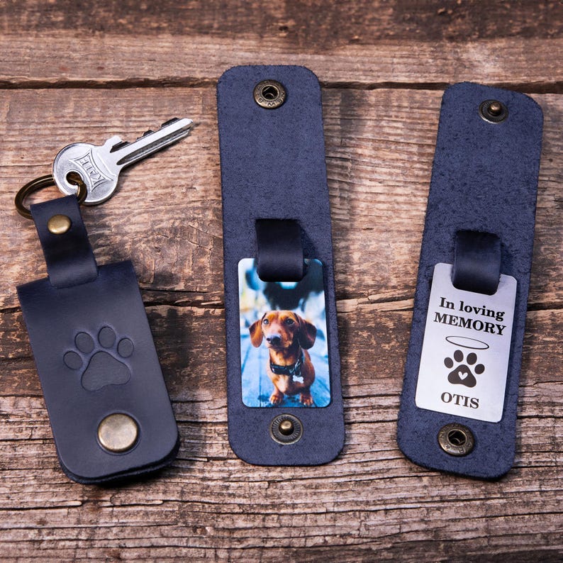 May include: Three dark blue leather key organizers with metal snaps. One features a paw print embossment and a silver key. Another displays a photo of a brown dachshund. The third shows a metal tag with the text In loving MEMORY, a halo, a paw print, and OTIS.