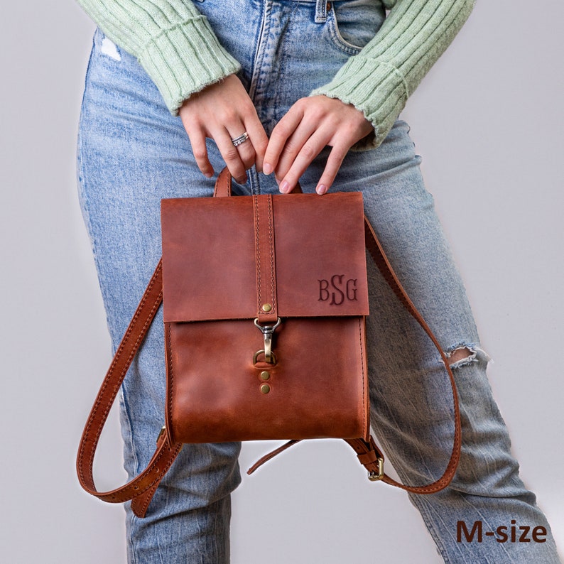 May include: A medium-sized brown leather backpack featuring a fold-over flap with a brass-toned metal clasp closure. The front of the bag is embossed with the monogram BSG. The backpack includes adjustable leather shoulder straps and brass rivets.