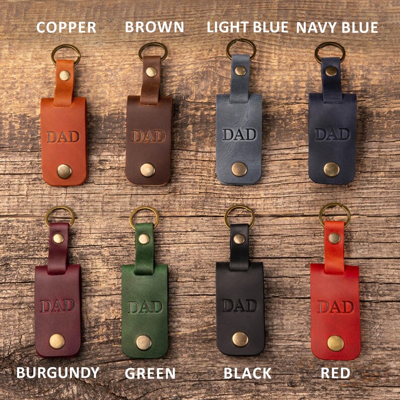 May include: Eight leather keychains arranged in two rows, each featuring a metal ring, a snap button, and the word DAD embossed on the front. The colors include copper, brown, light blue, navy blue, burgundy, green, black, and red leather.