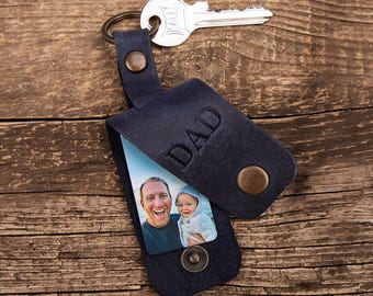May include: A dark blue leather keyring with a metal ring and a silver key attached. The leather flap is embossed with the word DAD and features a snap button closure. Inside, a rectangular photo shows an adult and a child smiling.