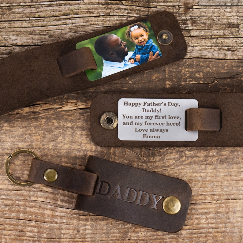 May include: Three dark brown leather keychains with brass-toned metal snaps. One keychain features a color photograph of a man and a child, another displays the engraved text: Happy Father's Day, Daddy! You are my first love, and my forever hero! Love always Emma, and the third is embossed with the word DADDY.