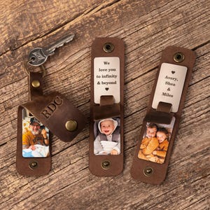 May include: Three brown leather keychains with metal snaps. One keychain holds a silver key and features the letters RDC and a photo of a person holding a baby. The others display metal plates with the text We love you to infinity & beyond or Avery, Shea & Miles, alongside photos of a baby and two children.
