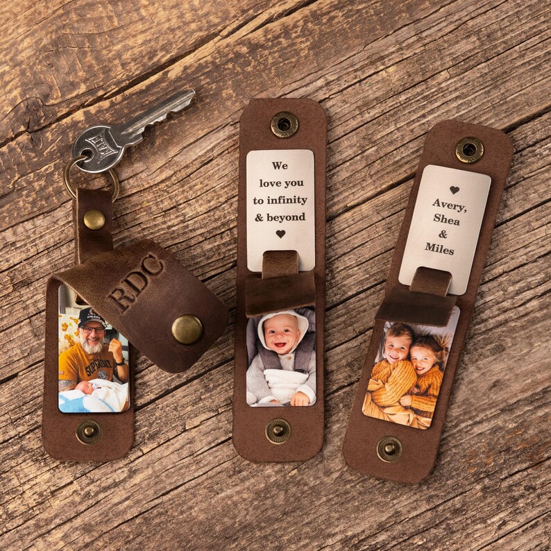 May include: Three brown leather keychains with metal snaps. One keychain holds a silver key and features the letters RDC and a photo of a person holding a baby. The others display metal plates with the text We love you to infinity & beyond or Avery, Shea & Miles, alongside photos of a baby and two children.