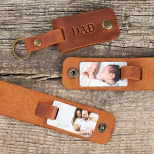 May include: Three brown leather keychains with brass hardware. One keychain is folded and embossed with the word DAD. The other two are unfolded, revealing rectangular photo inserts showing a sleeping infant and a family with a baby.