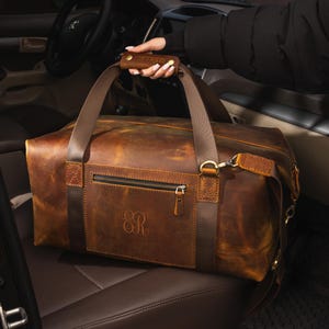 May include: A brown leather duffel bag featuring a horizontal zippered front pocket, dark brown leather straps, and a padded handle embossed with the name ROBERTS. The bag displays a monogram with the letters ER on the front and brass hardware.