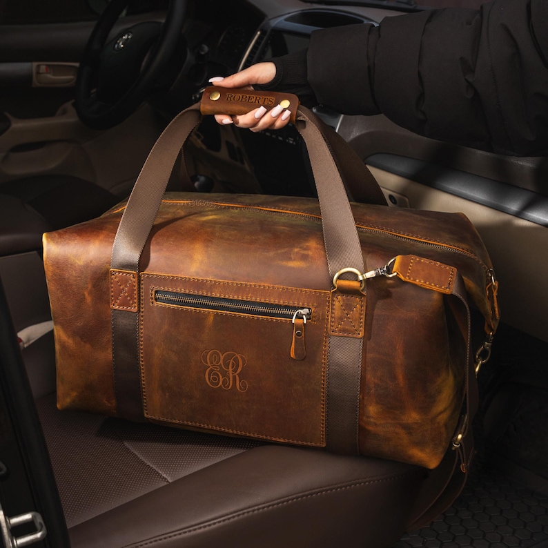 May include: A brown leather duffel bag featuring a horizontal zippered front pocket, dark brown leather straps, and a padded handle embossed with the name ROBERTS. The bag displays a monogram with the letters ER on the front and brass hardware.