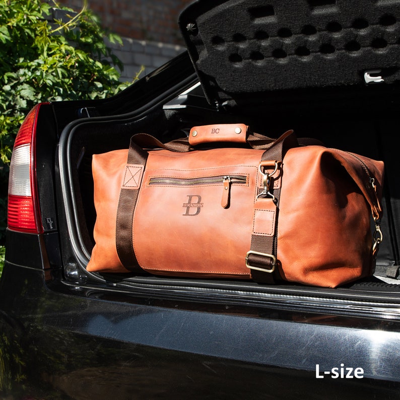 May include: A large tan leather duffel bag with brown nylon straps, a zippered front pocket, and brass hardware. The bag features a monogrammed handle with the letters BC and a large letter B embossed on the front. The text L-size is visible.