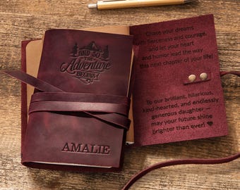 May include: A dark maroon leather journal with a wrap-around tie closure, featuring the embossed text "AND SO THE ADVENTURE BEGINS" and the name "AMALIE". The interior flap displays engraved text, and a gold-toned pen rests above the journal.