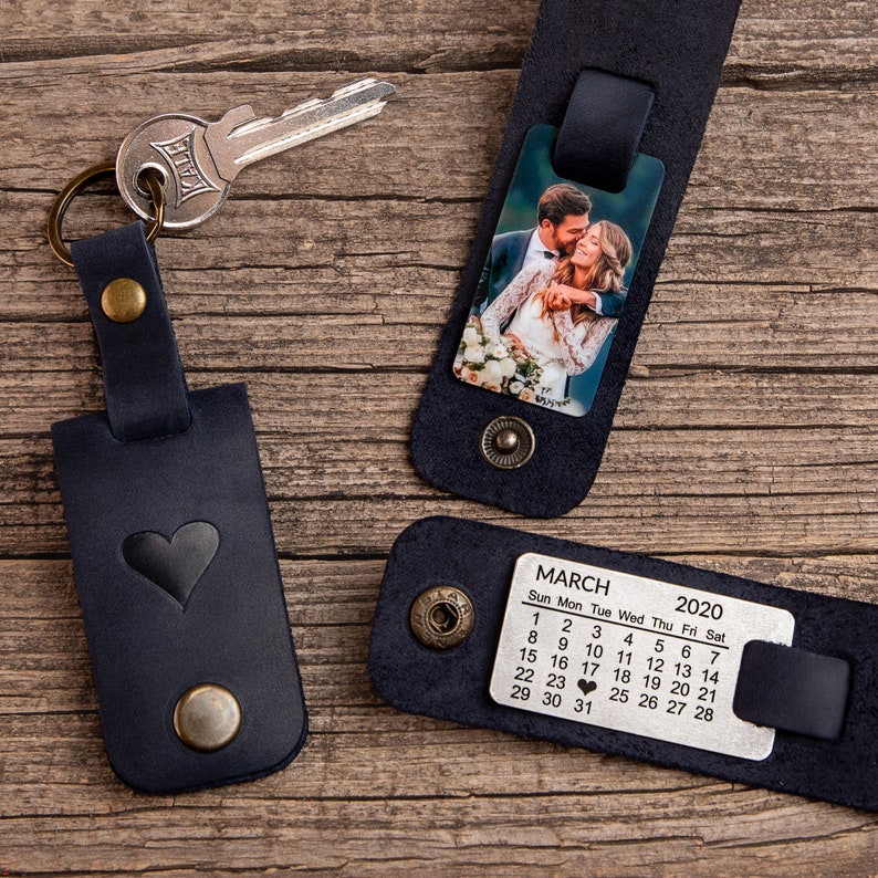 May include: Three dark blue leather keychains with brass hardware. One keychain holds a silver key, one displays a color photograph of a couple, and one features a metal plate engraved with a March 2020 calendar and a heart on the 24th.