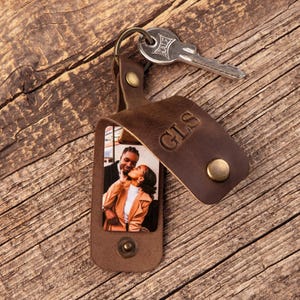 May include: A dark brown leather keychain featuring a fold-over flap secured with a brass snap button. The exterior is embossed with the letters GLS. Inside, a rectangular photo shows two people embracing. A silver metal key is attached to the ring.