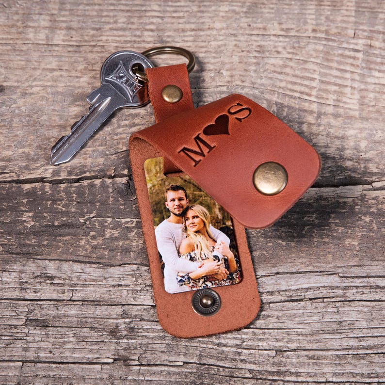 May include: A brown leather keychain with a metal ring and a silver key. The leather flap features the engraved letters M and S separated by a heart symbol. Beneath the flap, a rectangular photo shows a man and a woman embracing outdoors.